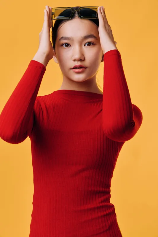 Young woman with smooth skin wearing red ribbed sweater and holding black sunglasses over head on yellow background. Portrait with confident gaze and casual pose in studio lighting