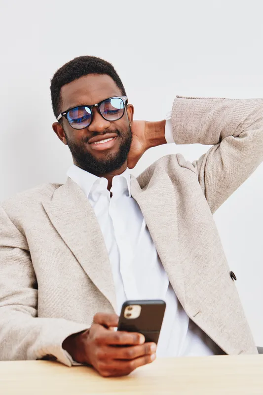 Confident smiling man of African descent wearing glasses, beige blazer and white shirt, holding smartphone, relaxed pose, indoor studio shot, casual lifestyle image.