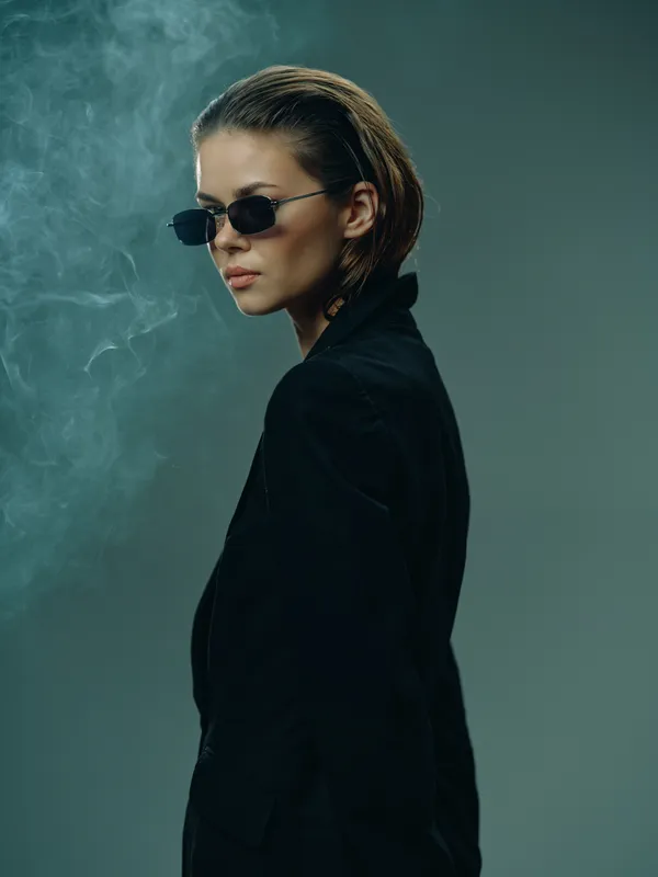 Young woman wearing sunglasses and black blazer with smoky background, stylish and confident expression.