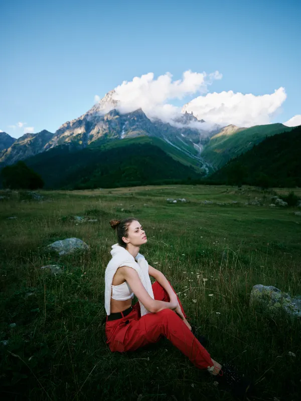 Woman sitting on a grassy meadow at the foot of impressive mountains, wearing red pants and a white knit vest, capturing a serene moment of outdoor nature and quiet travel contemplation