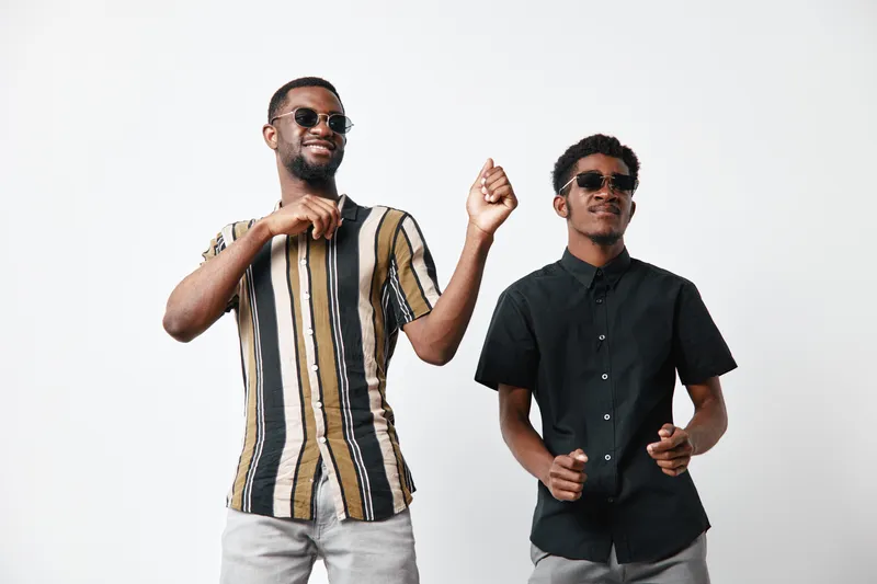 Two African American men with dark skin wearing sunglasses dancing and smiling in casual clothes against white background. Friends enjoying music and having fun together.