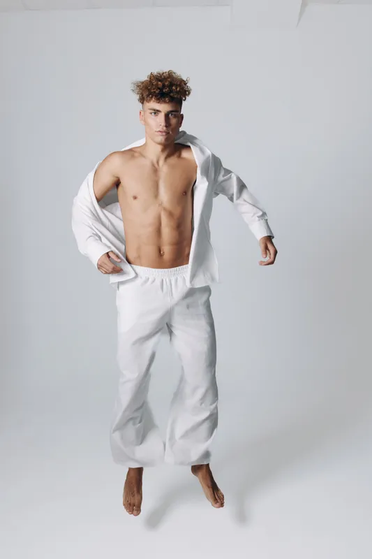 Young man with curly hair wearing white pants and open shirt jumping against plain background, showing athletic body and casual style in studio lighting.