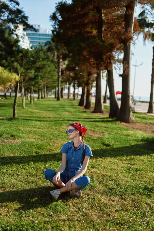 Lifestyle woman in modern boho-western denim street style sitting cross-legged on grass with sunglasses and red headscarf, film color effect in sunny park setting.