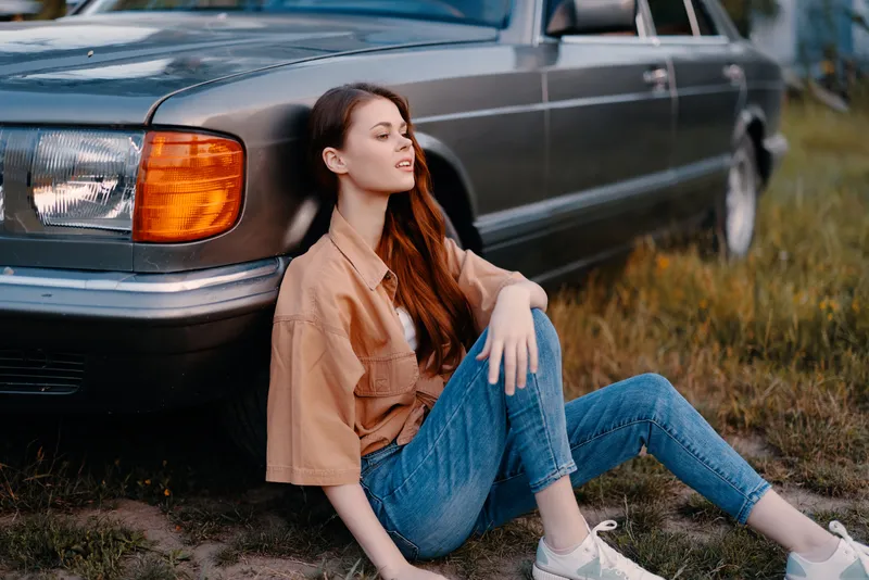 Young woman sitting by a vintage car, wearing a casual outfit, displaying a thoughtful expression amidst a natural backdrop, evoking a sense of nostalgia and freedom.