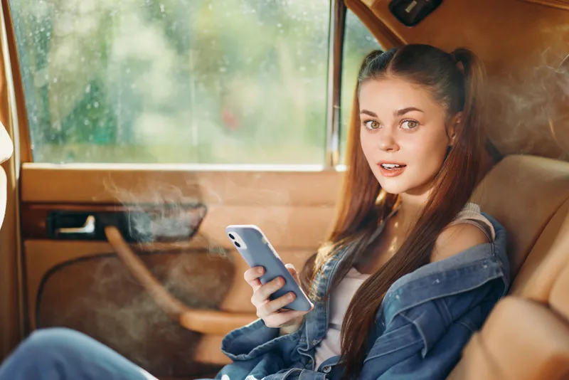 Young woman sitting in a vintage car, holding a smartphone, with casual denim attire and a cheerful expression, featuring a soft, natural background.