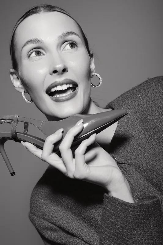 Happy Caucasian woman with light skin and earrings holds high heel shoe near face smiling in studio portrait.