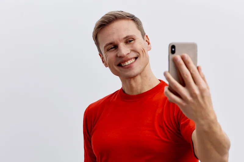 Smiling young man with short hair wearing bright red t-shirt takes a selfie with smartphone, isolated on a plain light gray background, studio portrait, casual lifestyle concept, cheerful mood.