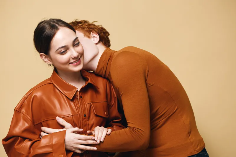 Couple in love with man kissing woman on cheek while she smiles, casual clothing and warm toned outfits in studio for romance and affection.