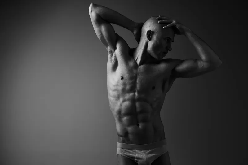 A muscular man poses dramatically in a striking black and white image, showcasing his toned physique and captivating expression in a minimalist setting.