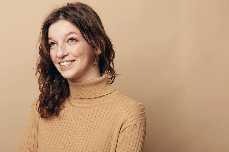 Smiling young woman with curly brown hair wearing a beige turtleneck sweater looks up with a happy expression on a plain beige background. Concept of joy and casual fashion.