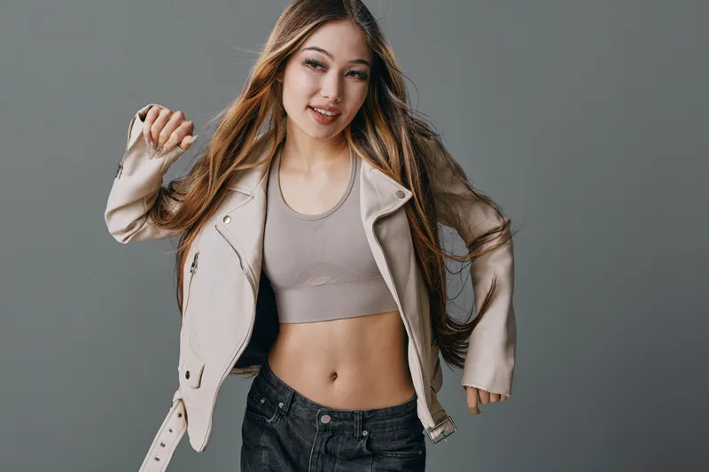 Young woman with long hair wearing a light jacket and crop top dancing on gray background. Casual style, smiling, hair flowing and motion captured in studio for fashion portrait.