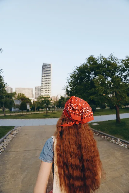 Lifestyle woman with long red hair wearing modern boho-western denim street style and a bright red bandana, captured with warm film color tones in an urban park setting.