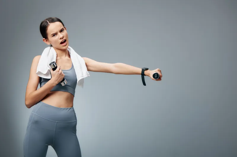 Fitness woman in gym attire performs a dynamic workout, holding a dumbbell with a towel over her shoulder, striking a confident athletic pose in a neutral studio.