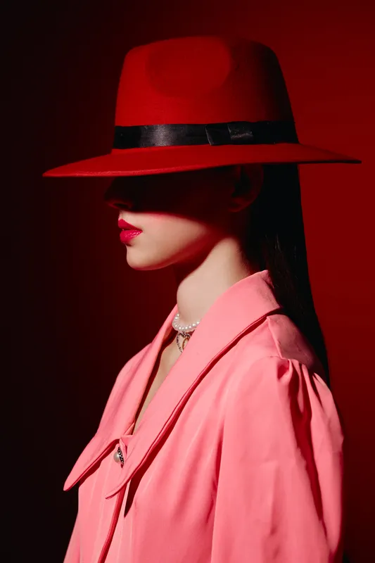 Young woman in vibrant red wide-brimmed hat casts shadow over her eyes. She wears elegant pink coat and pearl necklace in creative fashion portrait style. Beauty and style concept.