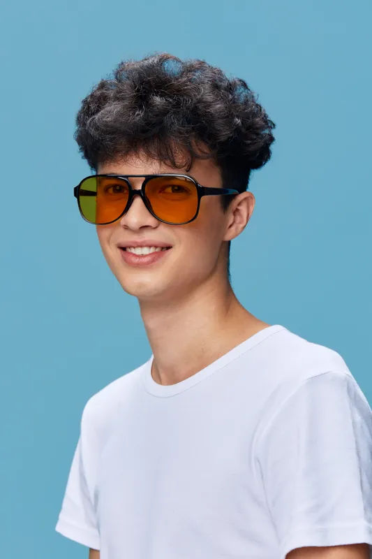 young man wearing orange sunglasses and white t-shirt smiling against blue background, curly hair, casual style, vibrant color contrast