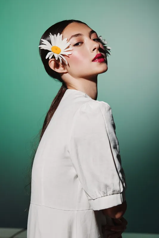 Elegant young woman with daisy flowers clipping her hair poses in a white dress against a green background. Bold makeup contrasts with soft petals. Creative fashion beauty concept.
