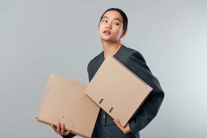 Woman in formal suit holding cardboard folders for documents looking away with thoughtful expression on neutral background in modern office or studio setting.