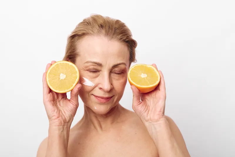 Mature woman with closed eyes holds two orange halves near face, applying cream on cheek. Concept of natural skincare, healthy lifestyle, and facial rejuvenation with citrus fruits.