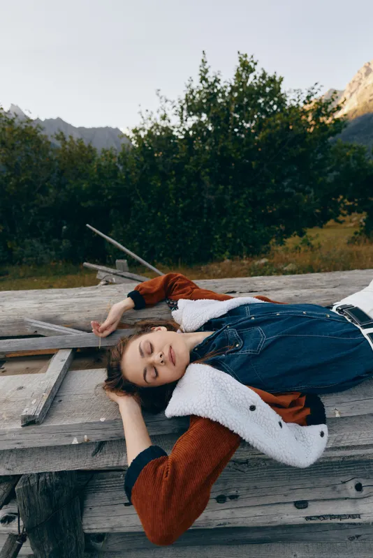 Woman lying outdoors on a wooden bench in countryside nature, a peaceful portrait of relaxation with closed eyes, casual jacket and mountains and forest scenery in soft sunlight.