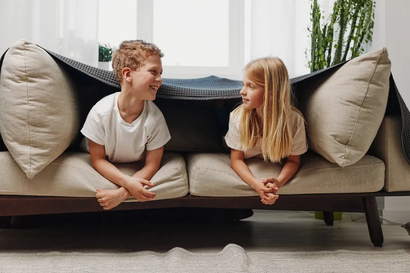 Two happy children lying on beige sofa facing each other smiling, cozy living room with natural light and neutral decor, concept of friendship and childhood joy.