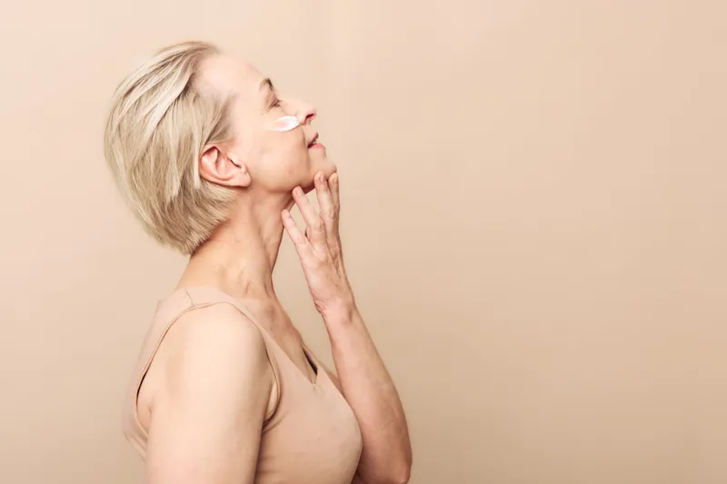 Mature woman with short blonde hair applies moisturizing cream on face in beige outfit. Soft lighting and neutral background emphasize skincare and natural beauty concept.