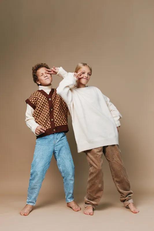 Two playful children pose against a warm beige backdrop, smiling and striking casual poses with knitwear and denim, capturing carefree childhood energy and charm.