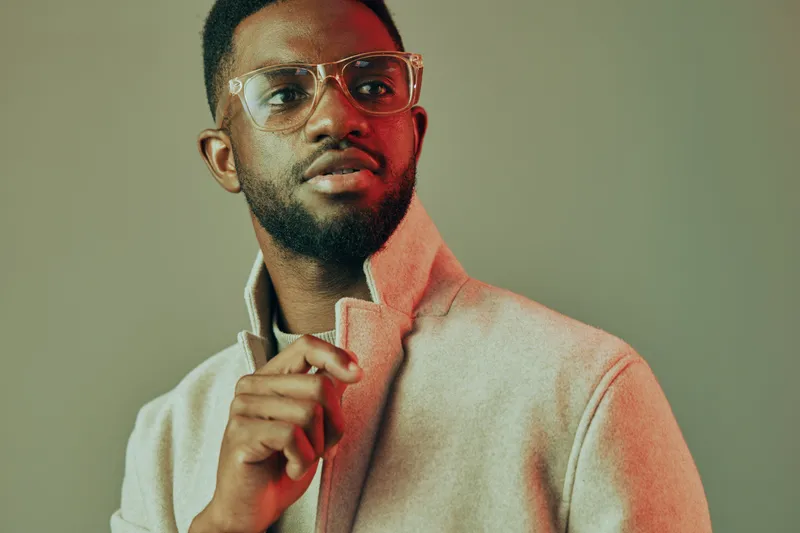 Stylish young African man in glasses wearing a modern grey jacket, showing confidence with a slight smile against a minimalistic background