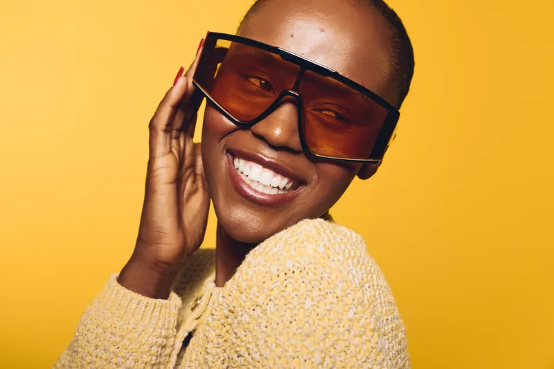 Smiling dark skinned African American woman wearing large brown sunglasses and yellow knitted sweater poses happily on yellow background. Concept of fashion, joy, style, and beauty.