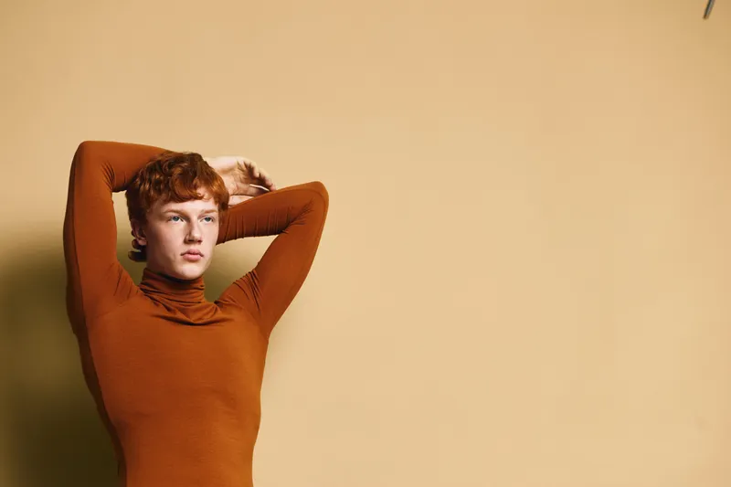 Young person with red curly hair in brown turtleneck standing against beige background with arms raised behind head, looking sideways with calm expression.