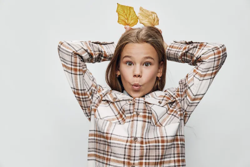 Girl with surprised expression wearing plaid shirt holding yellow autumn leaves behind head on light background. Child with fall leaves playing and showing emotion indoors.