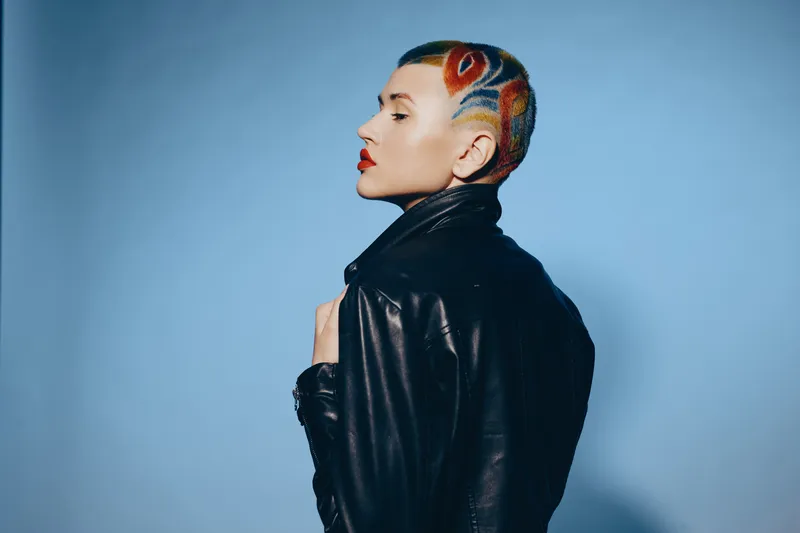 Woman with colorful shaved hairstyle and red lipstick wearing black leather jacket on blue background, expressing bold and artistic style confidently.