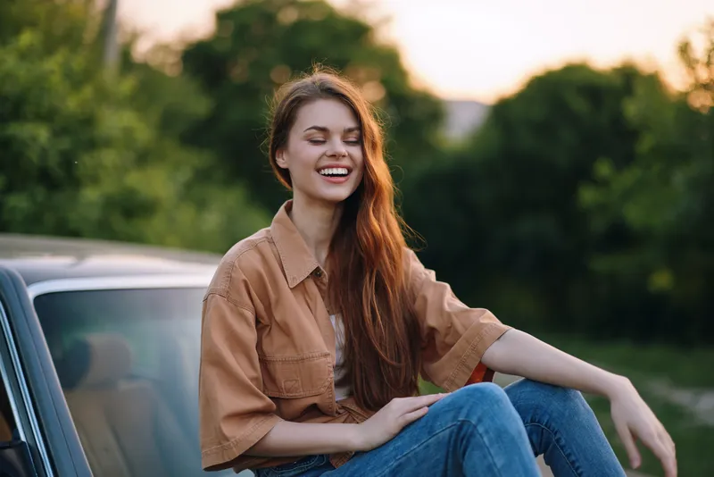 Happy young woman enjoying summer evening outdoors, wearing casual brown shirt and jeans, sitting on car hood, with greenery in the background and soft sunlight.
