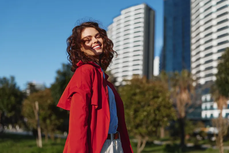 Smiling woman with curly hair wearing a red jacket in an outdoor urban park, modern buildings in the background. Bright, energetic portrait mood.
