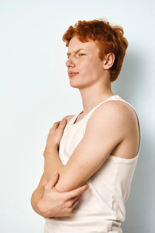 Young man with red hair in white sleeveless shirt looking sideways with skeptical expression isolated on white background. Portrait of thoughtful person in casual clothing.