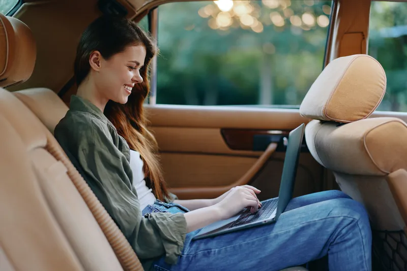 Stylish young woman working on a laptop in a car, enjoying the summer afternoon light with a joyful expression, reflecting a modern, mobile lifestyle.