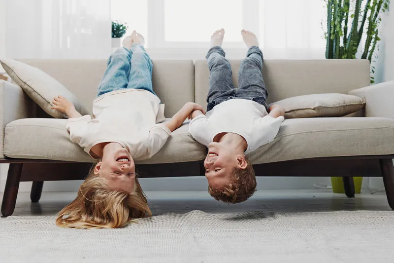 Two happy children lie upside down on a beige sofa in a bright living room, smiling and enjoying playful moments together. Concept of childhood fun and friendship.