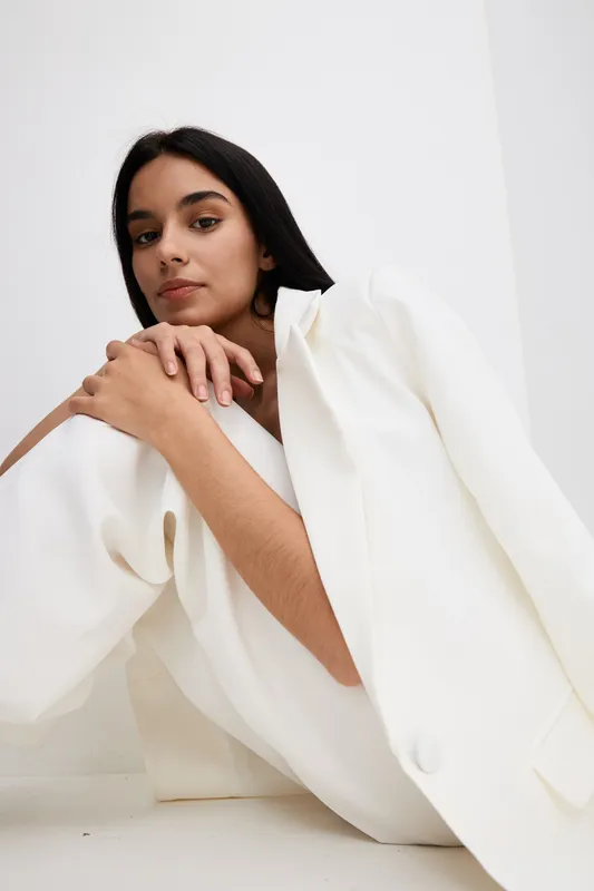 Young woman with dark hair wearing loose white suit sitting against minimalistic white background, showing calm expression and modern fashion style in natural light.