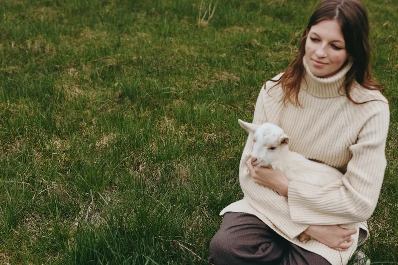 Woman in a cream sweater holds a white goat kid in a grassy field, intimate outdoor moment, wholesome lifestyle, calm care, warm natural light.