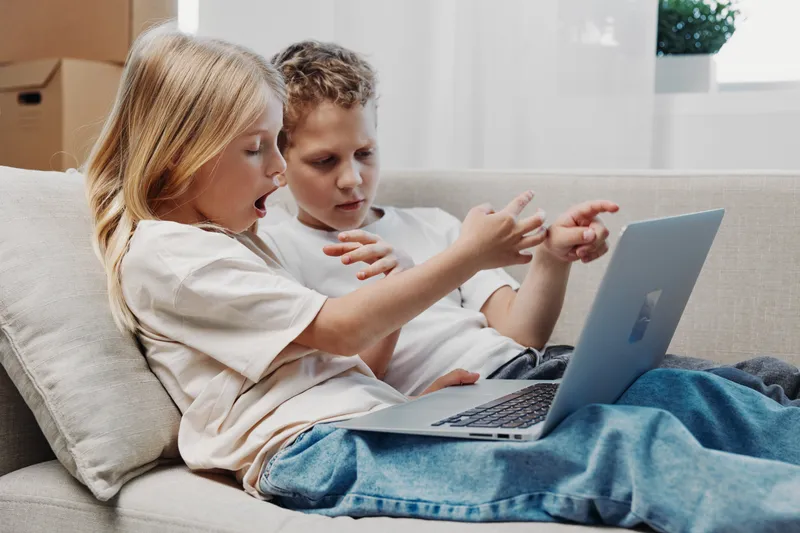 Two curious children sitting on couch using laptop together, pointing at screen with interest. Casual home environment with natural light and cozy atmosphere.