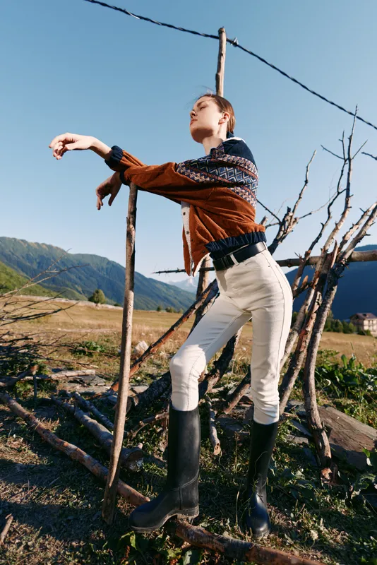 Woman model in countryside rural setting wearing fashion sweater and boots, elegant pose in field with mountains, rustic fence and sunlight for outdoor lifestyle portrait.