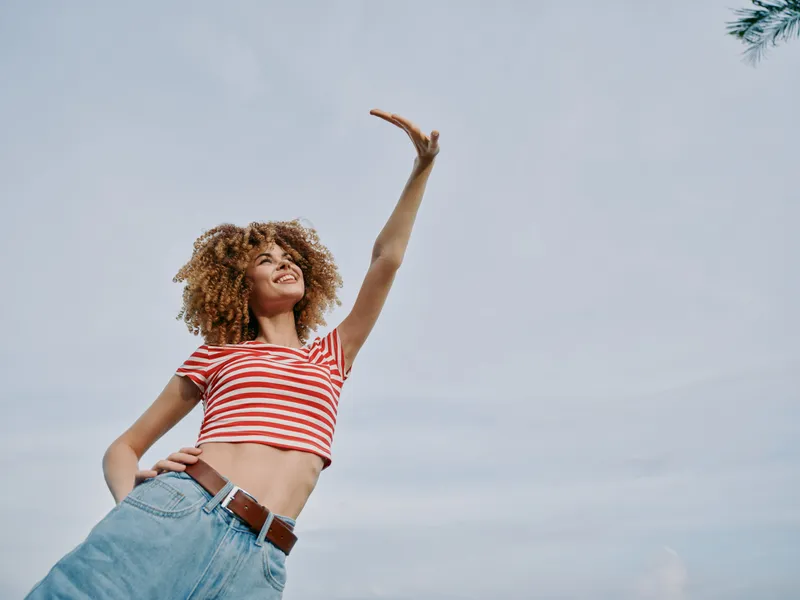 A joyful young woman with curly hair in a red striped crop top and denim jeans raises her arm toward the bright sky, capturing carefree energy and sunny optimism.