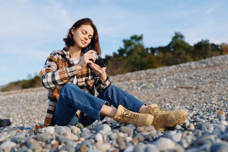 Woman sitting on a pebble beach, outdoors, holding a camera for photography, wearing a plaid shirt and jeans with boots, focused on a shot under clear sky and natural light