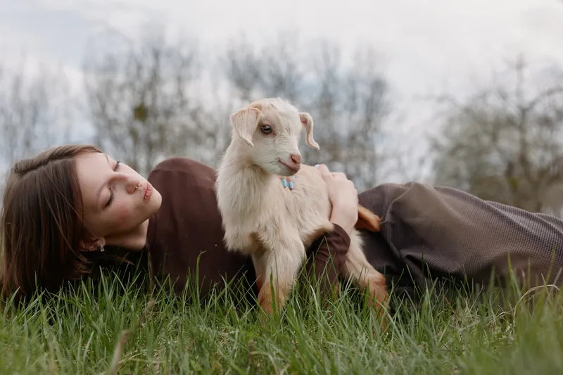 A woman lies on lush grass outdoors, gently cuddling a young goat kid, sharing a peaceful moment of friendship and affection in a bright natural field.
