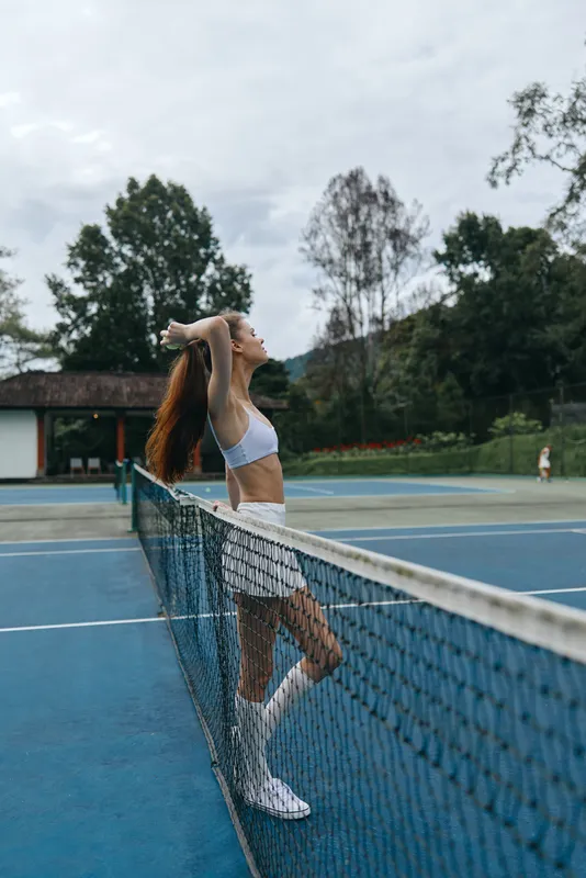 Young athletic woman in sportswear stretching by the tennis net, showcasing a fit physique and joyful expression against a vibrant outdoor background