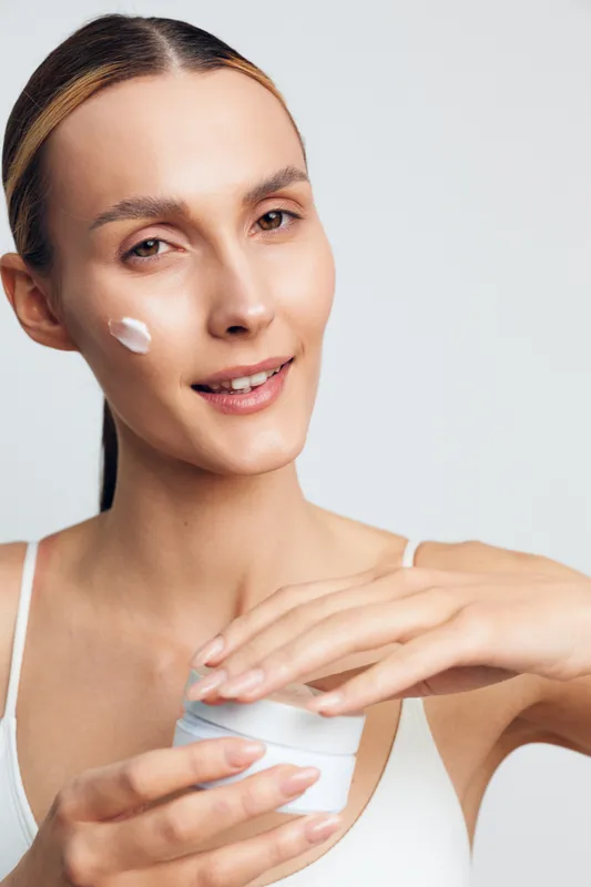 Young woman applying face cream with gentle smile, clean skin, natural makeup, soft lighting, white background, skincare routine, beauty care, moisturizing skin treatment.