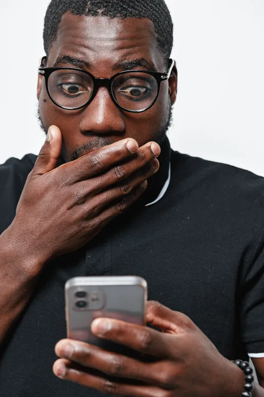 Black man with dark skin and glasses looking surprised at smartphone screen, covering mouth with hand in casual black shirt on white background.