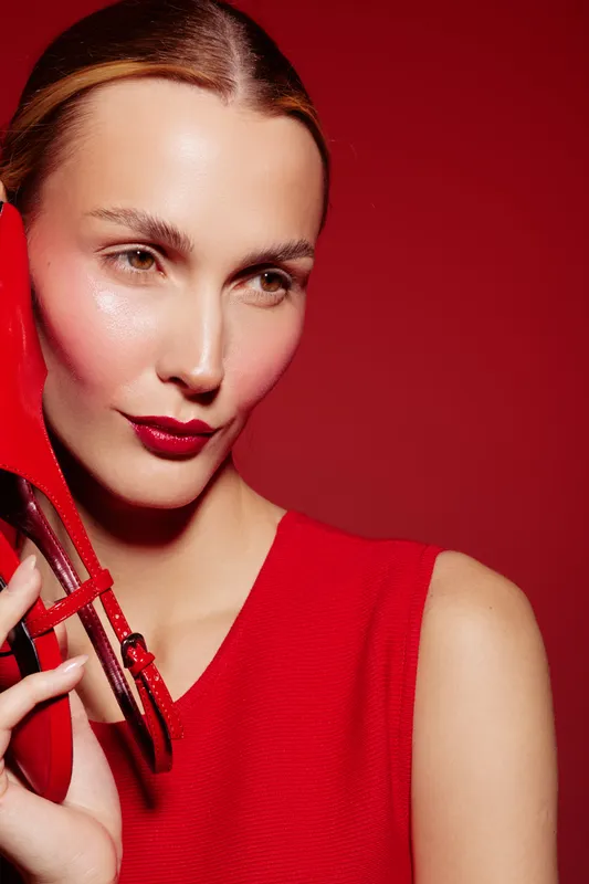 Close-up of a stylish woman in red dress holding a red high heel shoe near her face against a red background. Concept of fashion, elegance, beauty, and glamour.