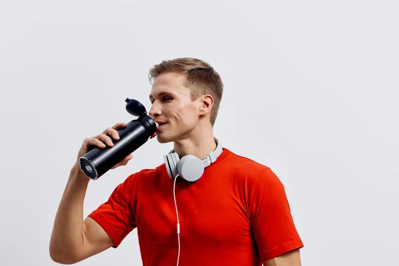 Smiling young man holding a black water bottle and wearing a red t-shirt with white headphones around his neck, looking to the side. Isolated on plain white background, casual lifestyle concept.