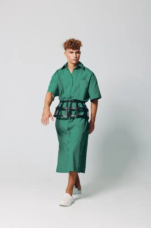 Young man walking in green shirt dress with black belt and white sneakers on plain background. Fashion model posing in casual outfit with curly hair and confident expression.