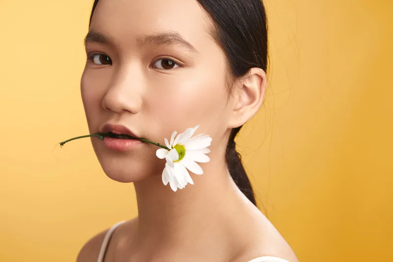 asian woman with a flower held gently in her lips smiles softly in a warm studio setting, radiating calm beauty and graceful presence.